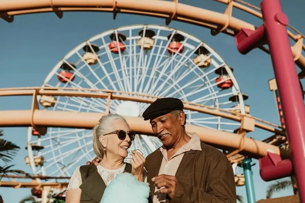 An older couple are standing in front of a carousel, at an amusement park. They are smiling and sharing cotton candy as they are now protected by life insurance with no medical exam.